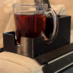 Puede incluir: Una bandeja negra para el brazo del sof&aacute;, impresa en 3D, con una taza de vidrio transparente llena de un l&iacute;quido oscuro. La taza tiene un asa y se encuentra en un compartimento de la bandeja. Un mando a distancia se encuentra detr&aacute;s de la taza.