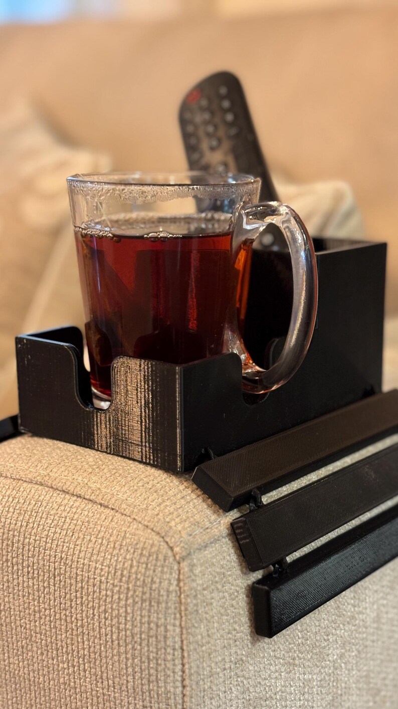 Puede incluir: Una bandeja negra para el brazo del sof&aacute;, impresa en 3D, con una taza de vidrio transparente llena de un l&iacute;quido oscuro. La taza tiene un asa y se encuentra en un compartimento de la bandeja. Un mando a distancia se encuentra detr&aacute;s de la taza.