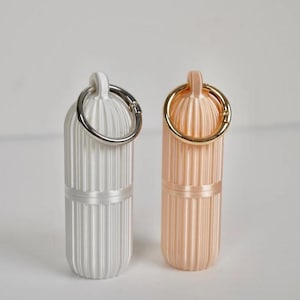 May include: Two cylindrical pill cases with vertical ribbed textures. One case is matte white with a silver-toned metal keyring attached to the top loop, and the other is a soft peach color with a gold-toned metal keyring attached to the top loop.