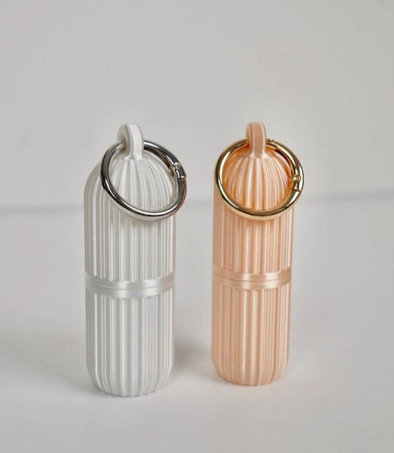 May include: Two cylindrical pill cases with vertical ribbed textures. One case is matte white with a silver-toned metal keyring attached to the top loop, and the other is a soft peach color with a gold-toned metal keyring attached to the top loop.