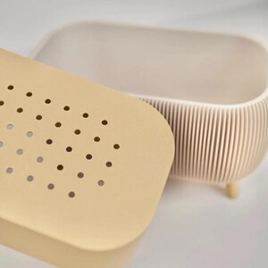 May include: A beige, oval-shaped container with a matching lid. The lid has a series of small, evenly spaced holes. The container's body has vertical ridges and rests on small, rounded feet. The design is minimalist and modern.
