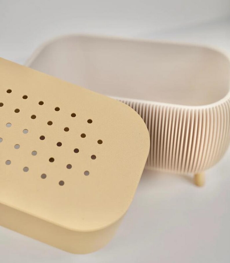 May include: A beige, oval-shaped container with a matching lid. The lid has a series of small, evenly spaced holes. The container's body has vertical ridges and rests on small, rounded feet. The design is minimalist and modern.