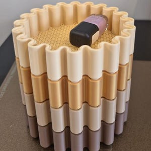May include: A multi-colored, cylindrical container with a wavy design. The container is composed of stacked layers in shades of beige, gold, and gray. A black and lavender cosmetic product rests on top.