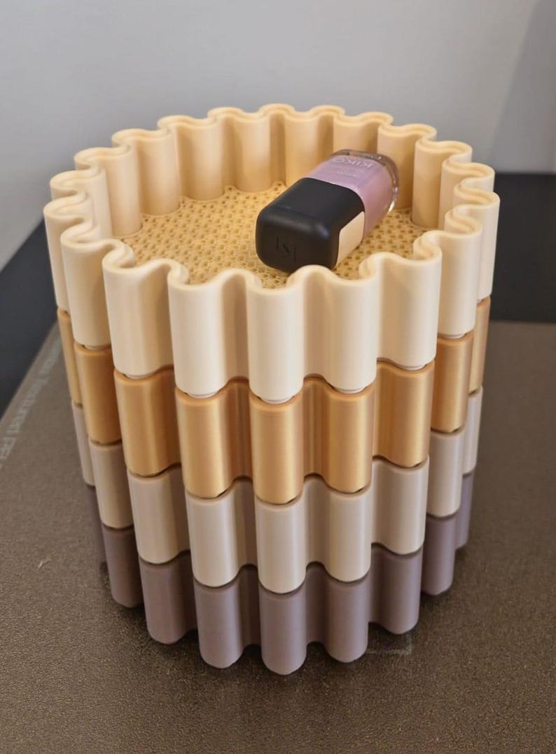 May include: A multi-colored, cylindrical container with a wavy design. The container is composed of stacked layers in shades of beige, gold, and gray. A black and lavender cosmetic product rests on top.