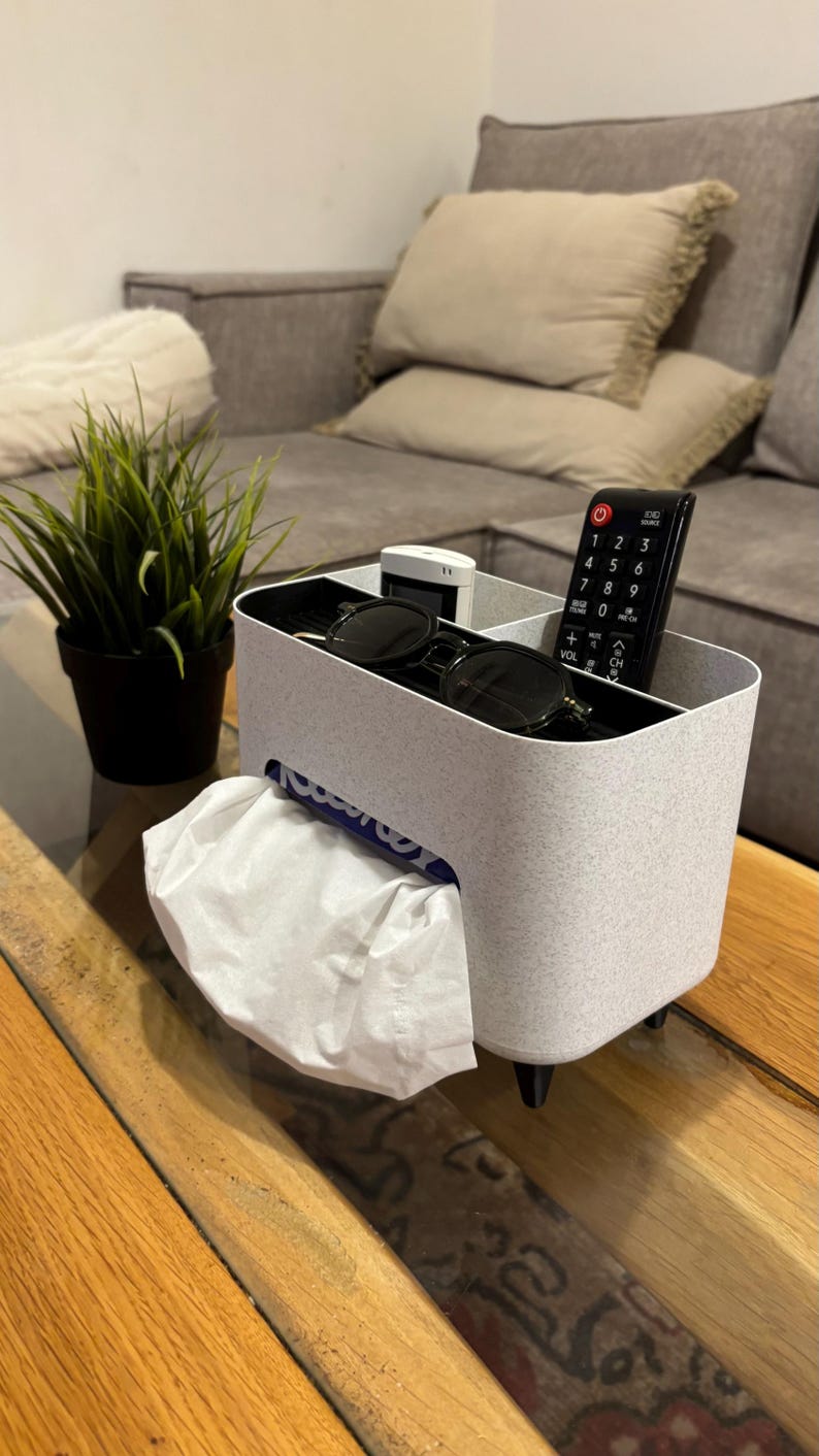 Op de afbeelding: Een lichtgrijze organizer met meerdere compartimenten met een tissue dispenser, zonnebril en een afstandsbediening. De organizer staat op een houten tafel. Links van de organizer staat een kleine plant in een pot.
