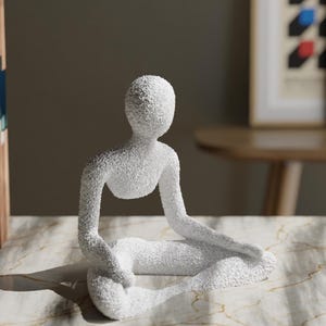 May include: A white, textured sculpture of a person in a seated yoga pose. The figure has a minimalist design and is placed on a marble-patterned surface. The sculpture is a decorative item.