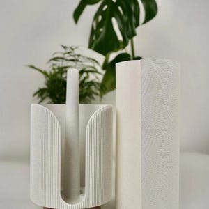 May include: A white paper towel holder with a matching roll of paper towels. The holder has a vertical design with a speckled texture and wooden feet. A green plant is in the background.