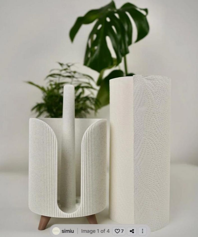 May include: A white paper towel holder with a matching roll of paper towels. The holder has a vertical design with a speckled texture and wooden feet. A green plant is in the background.