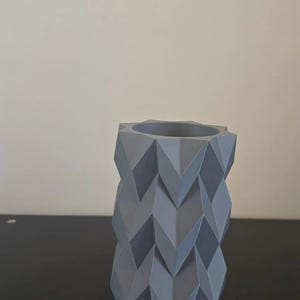 Puede incluir: Un jarr&oacute;n geom&eacute;trico gris con un patr&oacute;n en zigzag. El jarr&oacute;n es cil&iacute;ndrico con una abertura hexagonal en la parte superior. El jarr&oacute;n est&aacute; hecho de un material s&oacute;lido y se encuentra sobre una superficie oscura.
