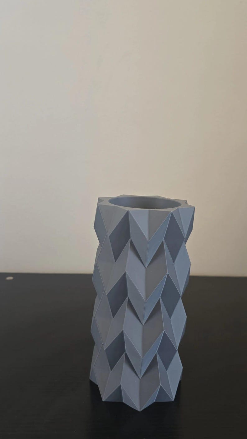Puede incluir: Un jarr&oacute;n geom&eacute;trico gris con un patr&oacute;n en zigzag. El jarr&oacute;n es cil&iacute;ndrico con una abertura hexagonal en la parte superior. El jarr&oacute;n est&aacute; hecho de un material s&oacute;lido y se encuentra sobre una superficie oscura.