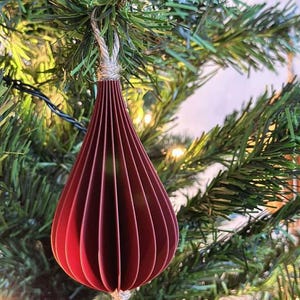 May include: A burgundy paper teardrop ornament hanging from a Christmas tree. The ornament is made of layered paper and has a textured, ribbed appearance. It is suspended by a natural twine string, and the tree's green needles and lights provide a festive backdrop.