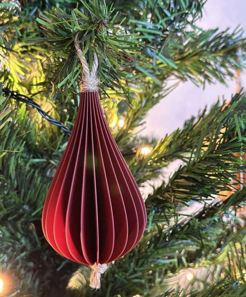 May include: A burgundy paper teardrop ornament hanging from a Christmas tree. The ornament is made of layered paper and has a textured, ribbed appearance. It is suspended by a natural twine string, and the tree's green needles and lights provide a festive backdrop.