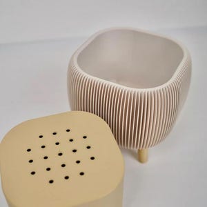 May include: A beige ceramic planter with a ridged exterior and a matching lid with small holes. The planter has a square top and rounded sides, supported by three small wooden legs. The lid sits beside the planter.