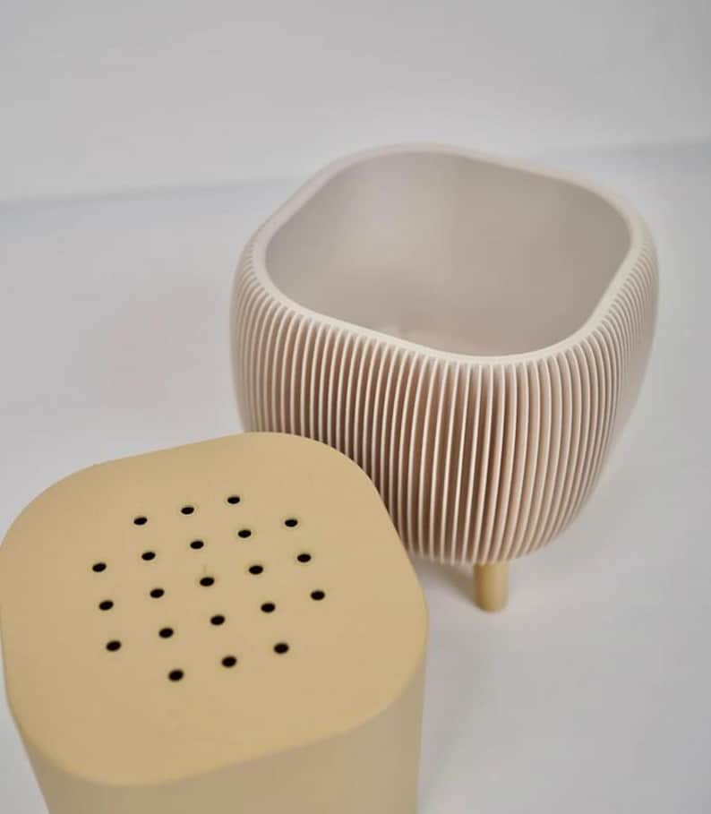 May include: A beige ceramic planter with a ridged exterior and a matching lid with small holes. The planter has a square top and rounded sides, supported by three small wooden legs. The lid sits beside the planter.