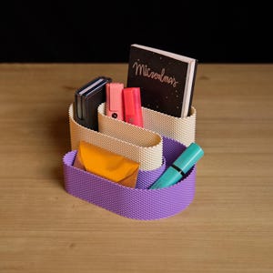 May include: A two-tiered desk organiser with a textured surface, featuring a cream-coloured upper section and a purple lower section. It holds a black notebook with the text "Microalmas", two pink highlighters, a dark wallet, an orange envelope, and a teal pen.