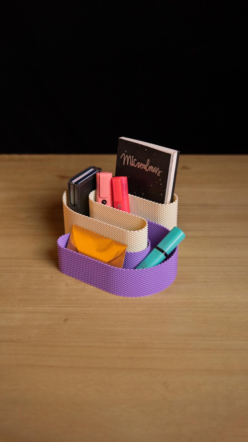 May include: A two-tiered desk organiser with a textured surface, featuring a cream-coloured upper section and a purple lower section. It holds a black notebook with the text "Microalmas", two pink highlighters, a dark wallet, an orange envelope, and a teal pen.