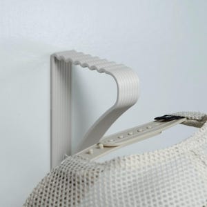 May include: A light gray hat rack mounted on a white wall, holding a mesh trucker hat. The rack has a wavy design and is made of a light gray material. The hat is a light beige color.