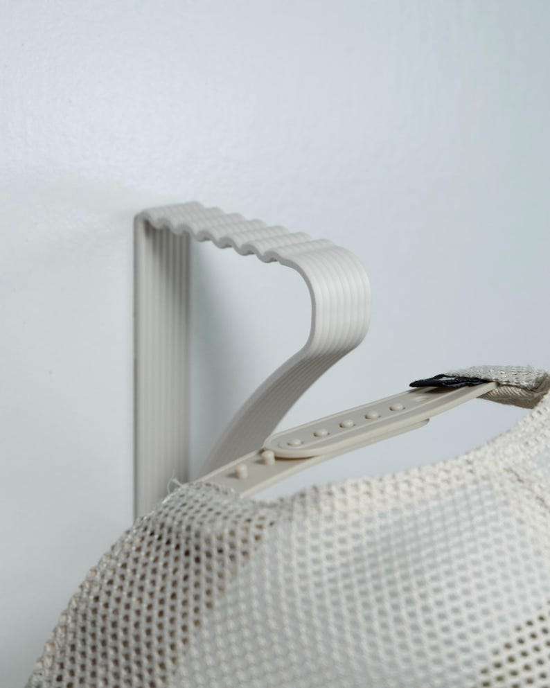 May include: A light gray hat rack mounted on a white wall, holding a mesh trucker hat. The rack has a wavy design and is made of a light gray material. The hat is a light beige color.