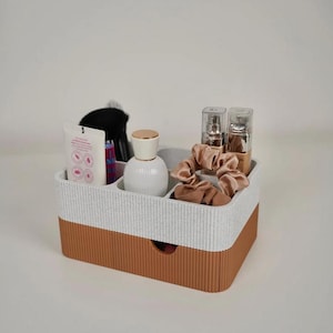 May include: A two-tone, textured organizer with a white and brown color scheme. The top section is divided into compartments holding beauty products, including a brush, perfume, and makeup. The bottom section has a small drawer.