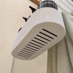K&ouml;nnte beinhalten: Ein cremefarbener, ovaler Duschkorb mit gerippter Au&szlig;enseite und einer Reihe paralleler Abflussschlitze am Boden. Der Korb enth&auml;lt schwarze Pumpflaschen, von denen eine deutlich mit den W&ouml;rtern SHAMPOO und SILK beschriftet ist.
