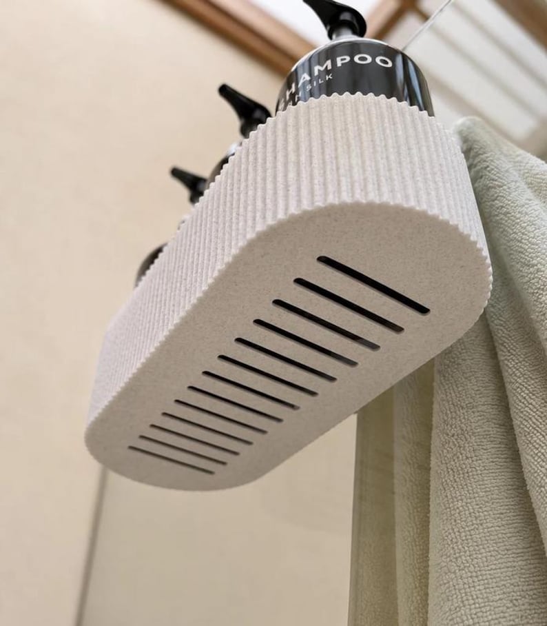 K&ouml;nnte beinhalten: Ein cremefarbener, ovaler Duschkorb mit gerippter Au&szlig;enseite und einer Reihe paralleler Abflussschlitze am Boden. Der Korb enth&auml;lt schwarze Pumpflaschen, von denen eine deutlich mit den W&ouml;rtern SHAMPOO und SILK beschriftet ist.