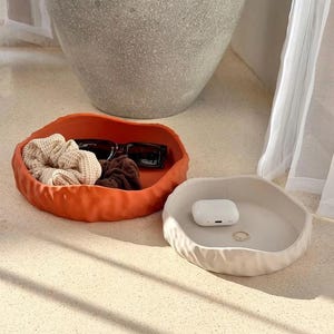 May include: Two round, shallow trays with irregular, wavy edges. One tray is a vibrant burnt orange color containing black sunglasses, a beige waffle-textured scrunchie, and a brown scrunchie. The other tray is cream-colored and holds white wireless earbuds and a small gold ring.