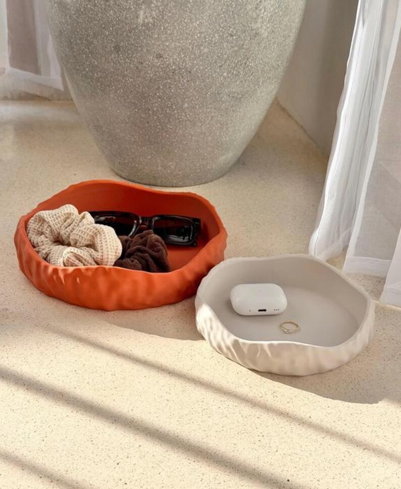 May include: Two round, shallow trays with irregular, wavy edges. One tray is a vibrant burnt orange color containing black sunglasses, a beige waffle-textured scrunchie, and a brown scrunchie. The other tray is cream-colored and holds white wireless earbuds and a small gold ring.