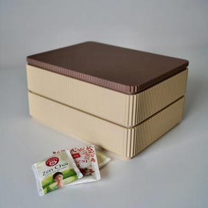 May include: A two-tiered beige box with a ribbed design and a brown lid. The box is rectangular and sits on a white surface. Tea packets with the text "Zen Chai" are in front of the box.