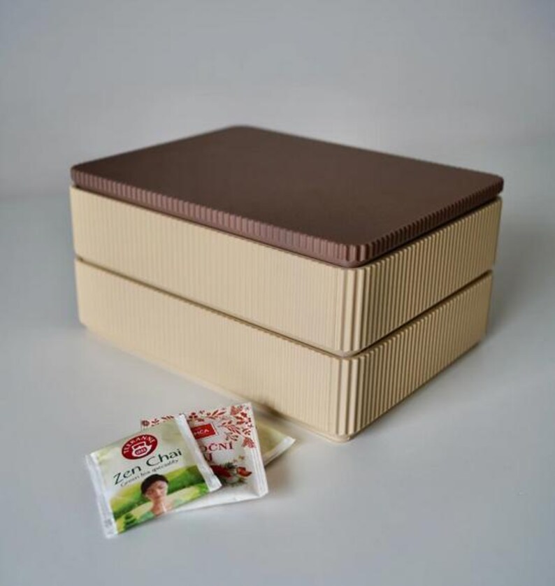 May include: A two-tiered beige box with a ribbed design and a brown lid. The box is rectangular and sits on a white surface. Tea packets with the text "Zen Chai" are in front of the box.