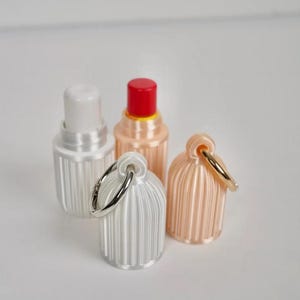 May include: Four small, cylindrical containers with vertical ribbed textures. Two containers are open, revealing white and red inner caps. Two containers are closed with attached silver-toned metal rings for hanging, one in white and one in a pale peach color.