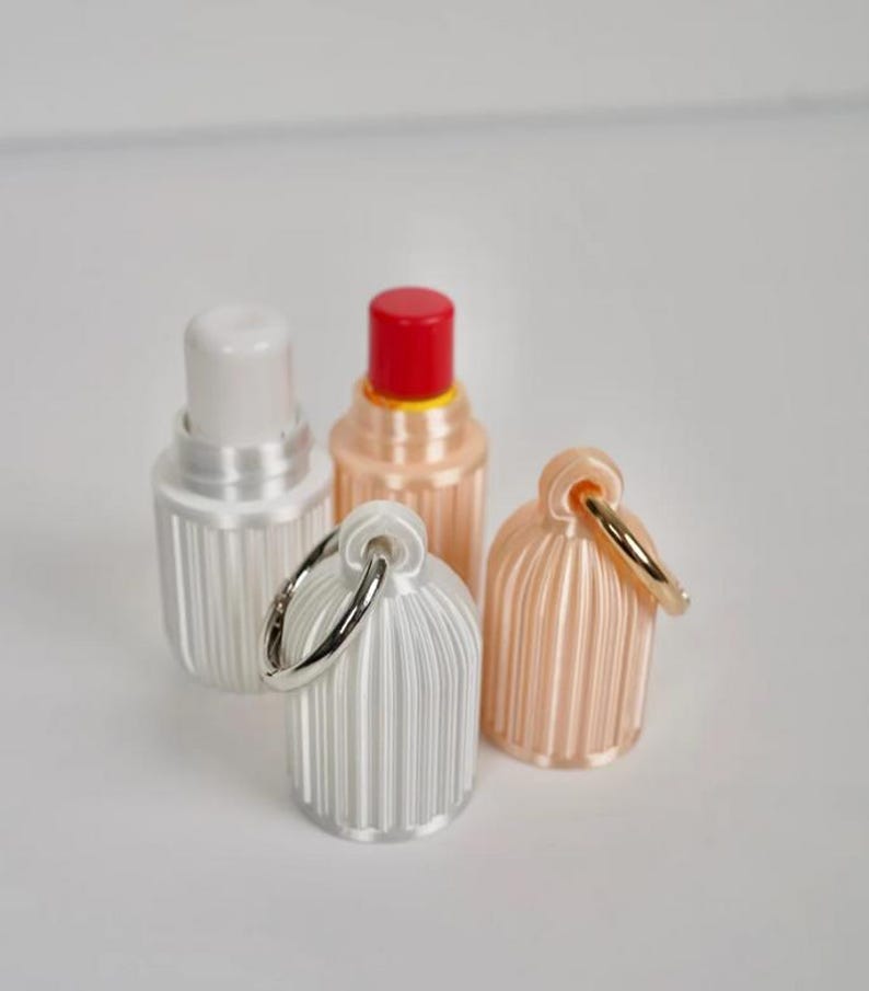 May include: Four small, cylindrical containers with vertical ribbed textures. Two containers are open, revealing white and red inner caps. Two containers are closed with attached silver-toned metal rings for hanging, one in white and one in a pale peach color.