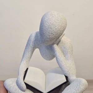May include: A white, textured sculpture of a person seated with legs crossed, reading a book. The figure's head rests on a hand, and the book is open. The sculpture has a minimalist design and a neutral colour palette.