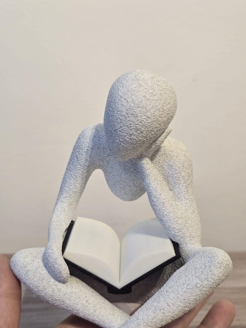 May include: A white, textured sculpture of a person seated with legs crossed, reading a book. The figure's head rests on a hand, and the book is open. The sculpture has a minimalist design and a neutral colour palette.