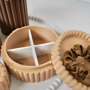 May include: A round, beige, scalloped-edge jewelry box with a removable white speckled cross-divider inside. The lid is detached and rests beside the base, containing a brown satin scrunchie. A gold chain is visible in one compartment.