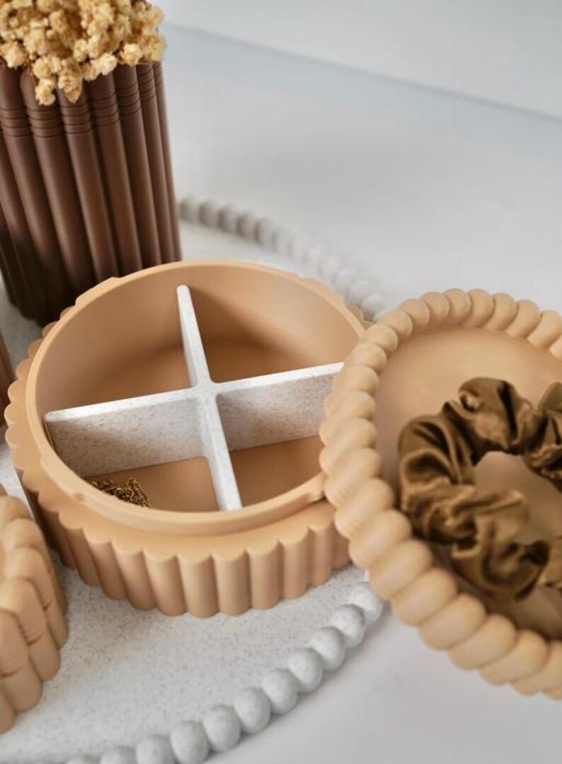 May include: A round, beige, scalloped-edge jewelry box with a removable white speckled cross-divider inside. The lid is detached and rests beside the base, containing a brown satin scrunchie. A gold chain is visible in one compartment.