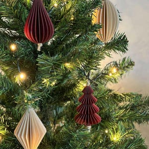 May include: Christmas tree ornaments in various shapes and colors. The ornaments include a teardrop, a tree, a diamond, and an oval. The ornaments are made of paper and are shades of red and beige. The ornaments are hanging on a green Christmas tree.