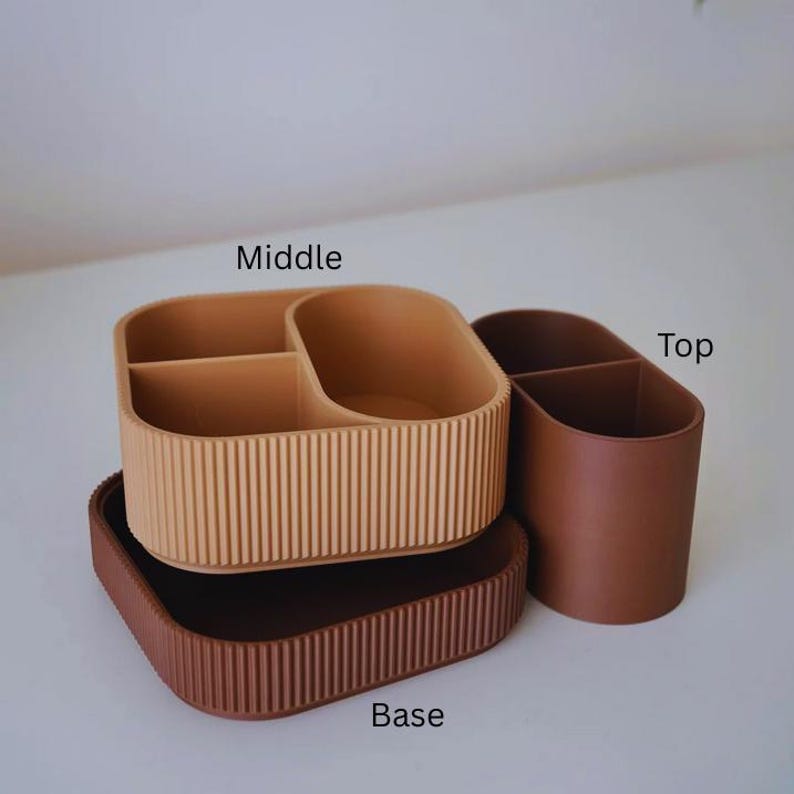 May include: A three-piece desk organizer set in shades of brown. The set includes a rectangular base, a divided middle section, and a single-compartment top piece. The middle section has vertical ribbed detailing, while the base has a similar design.