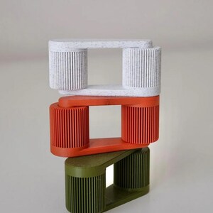 May include: Three stacked, geometric sculptures in white, orange, and olive green. Each sculpture has a rectangular top and bottom connected by two cylindrical, ribbed columns. The white sculpture has a speckled pattern.