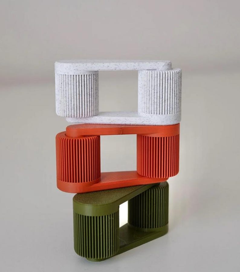 May include: Three stacked, geometric sculptures in white, orange, and olive green. Each sculpture has a rectangular top and bottom connected by two cylindrical, ribbed columns. The white sculpture has a speckled pattern.