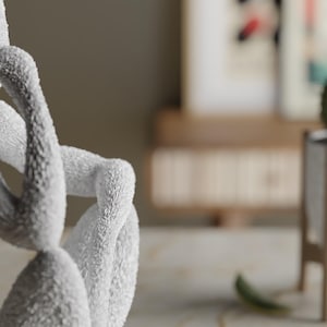 Puede incluir: Una escultura suave y blanca de una figura en una pose pensativa. Los brazos de la figura est&aacute;n cruzados y su cabeza descansa sobre sus manos. La escultura tiene una superficie texturizada. En el fondo, se ve un cactus en una maceta blanca.