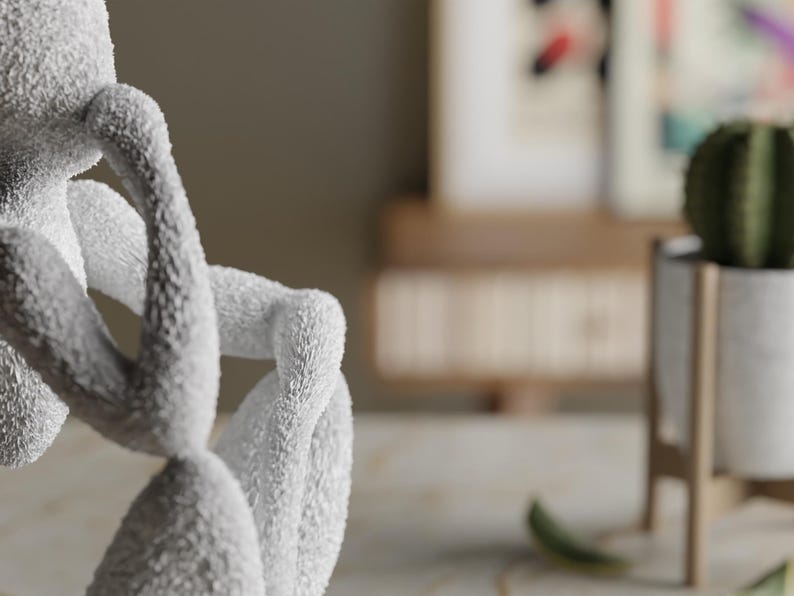 Puede incluir: Una escultura suave y blanca de una figura en una pose pensativa. Los brazos de la figura est&aacute;n cruzados y su cabeza descansa sobre sus manos. La escultura tiene una superficie texturizada. En el fondo, se ve un cactus en una maceta blanca.