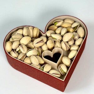 May include: A heart-shaped red box filled with pistachios. The box has a small heart-shaped cutout in the centre. The pistachios are in their shells, with some shells open, revealing the green nut inside. The box is on a white background.