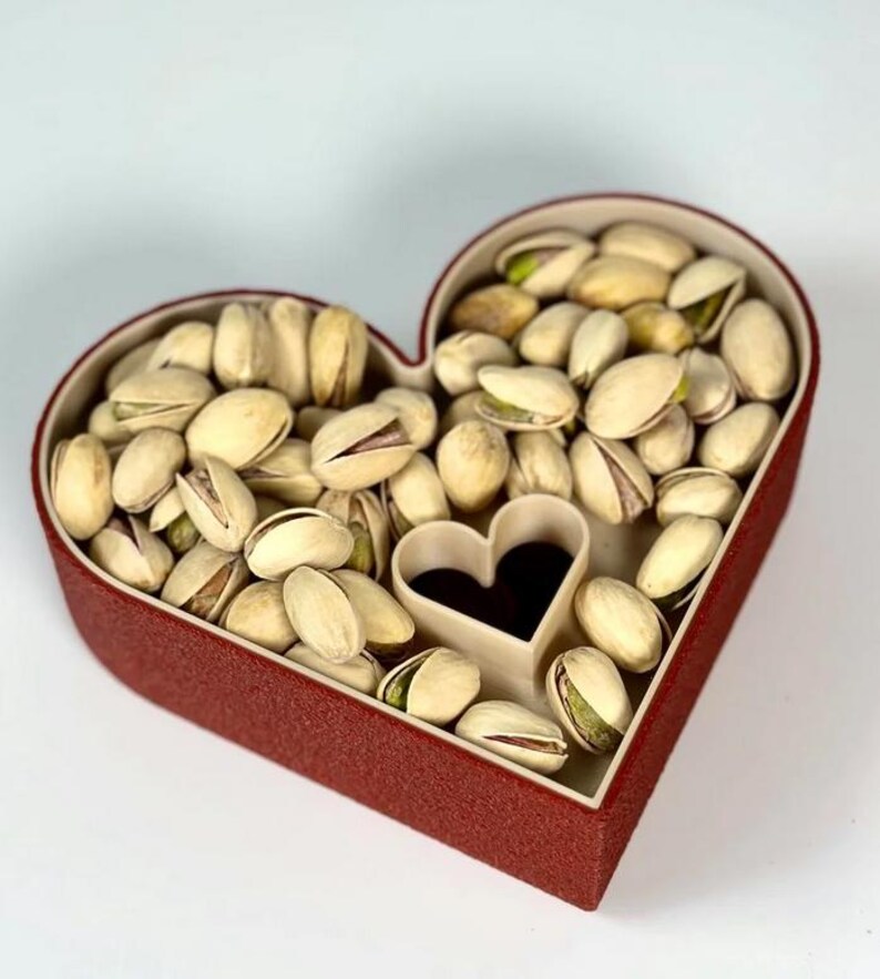 May include: A heart-shaped red box filled with pistachios. The box has a small heart-shaped cutout in the centre. The pistachios are in their shells, with some shells open, revealing the green nut inside. The box is on a white background.