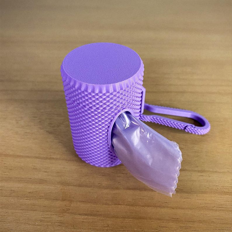 May include: A lavender-colored pet waste bag dispenser with a textured, woven-like design. The dispenser has a rounded top and a plastic clip for attachment. A partially visible bag is emerging from the dispenser.