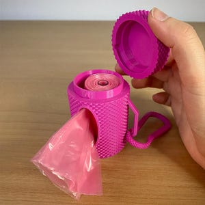 May include: A bright pink, 3D-printed dog waste bag dispenser. The dispenser has a textured surface, a handle, and a screw-on lid. A roll of pink bags is visible inside, with a bag extending from the side opening.