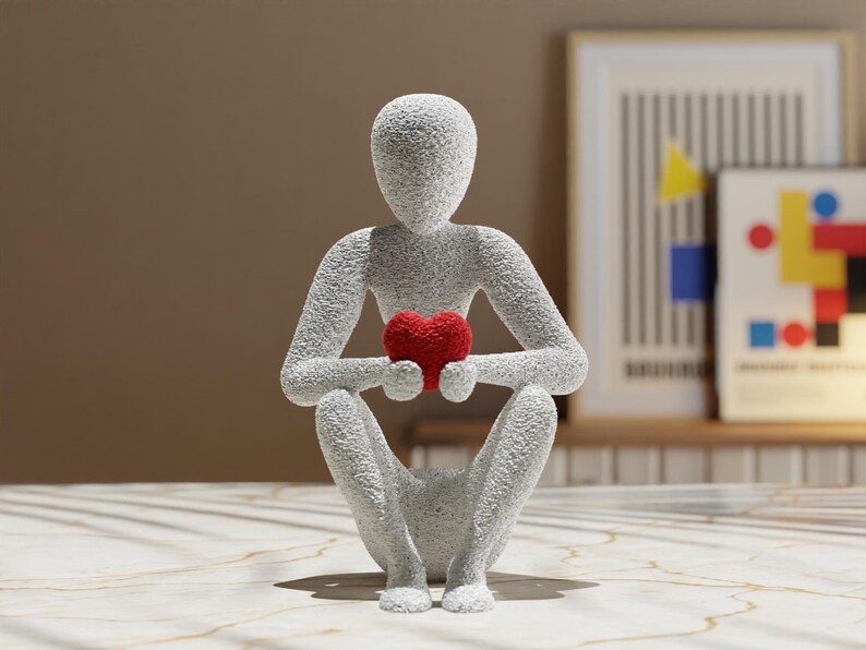 Puede incluir: Escultura gris texturizada de una persona sentada con las rodillas dobladas, sosteniendo un coraz&oacute;n rojo. La figura tiene una superficie rugosa y granular. Es un art&iacute;culo decorativo.