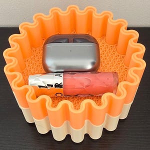 May include: An orange and white wavy-edged container holds a silver wireless earbud case and a tube of pink lip gloss. The container has a textured interior and is sitting on a dark surface.