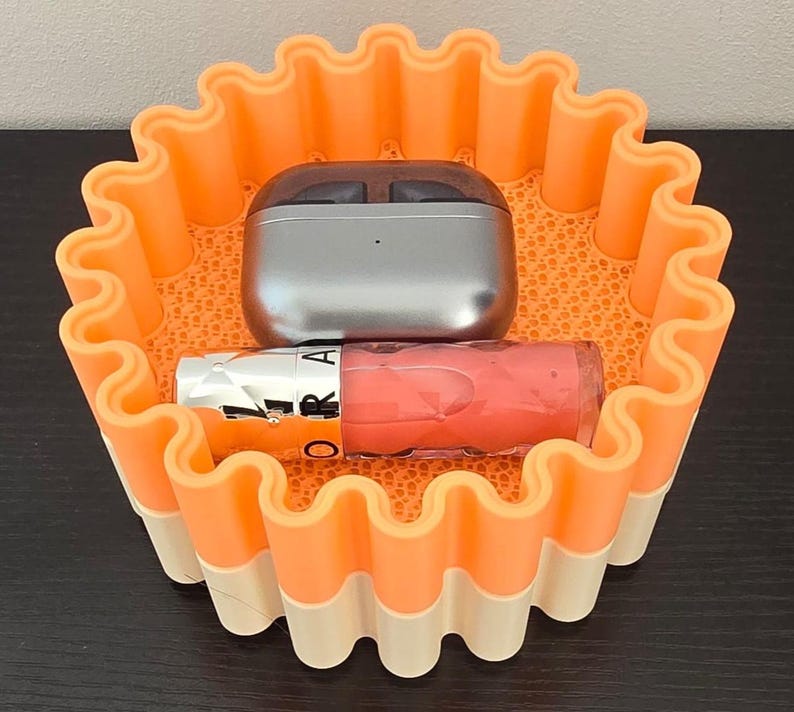 May include: An orange and white wavy-edged container holds a silver wireless earbud case and a tube of pink lip gloss. The container has a textured interior and is sitting on a dark surface.