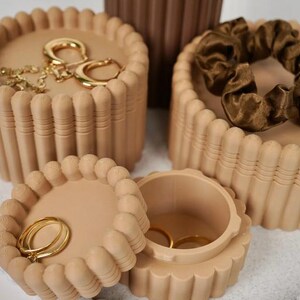 May include: Three round beige storage containers with ribbed sides and scalloped edges. One container is open, revealing two gold rings inside. Another container holds gold hoop earrings, while the third holds a dark brown fabric hair scrunchie.