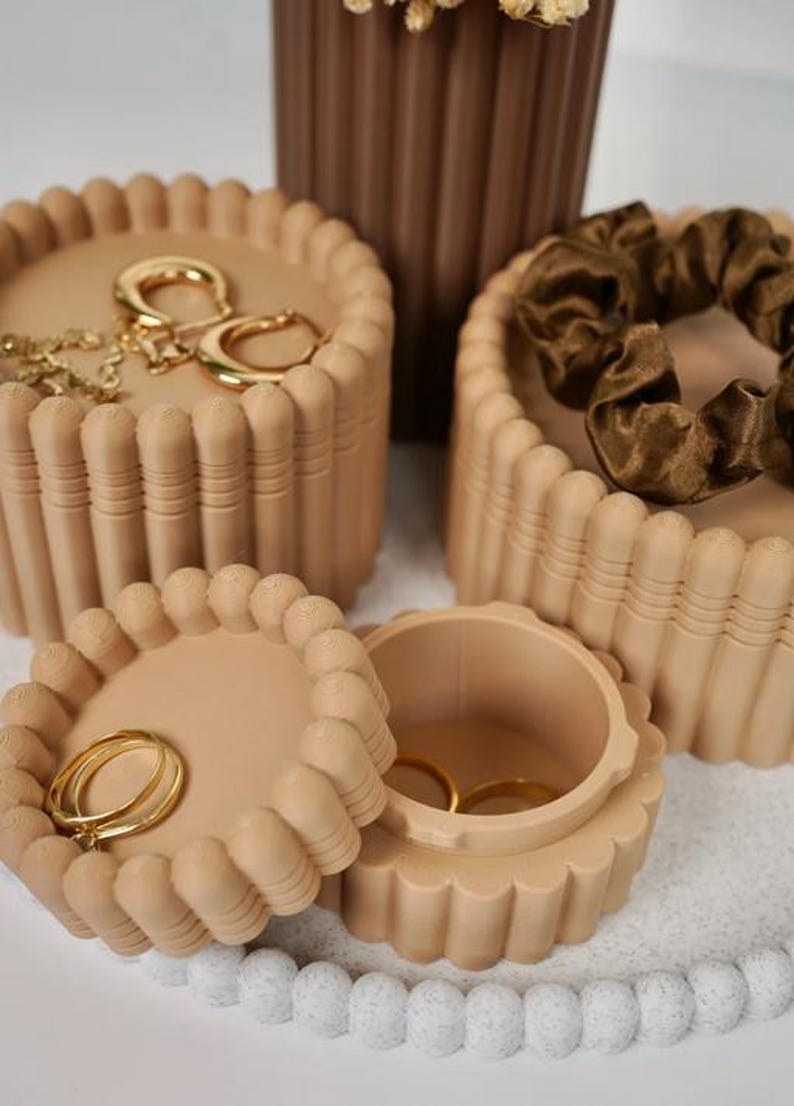 May include: Three round beige storage containers with ribbed sides and scalloped edges. One container is open, revealing two gold rings inside. Another container holds gold hoop earrings, while the third holds a dark brown fabric hair scrunchie.